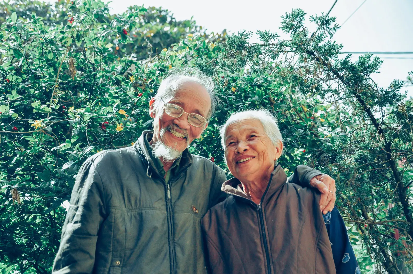 A smiling couple standing arm in arm beneath the trees on a bright afternoon