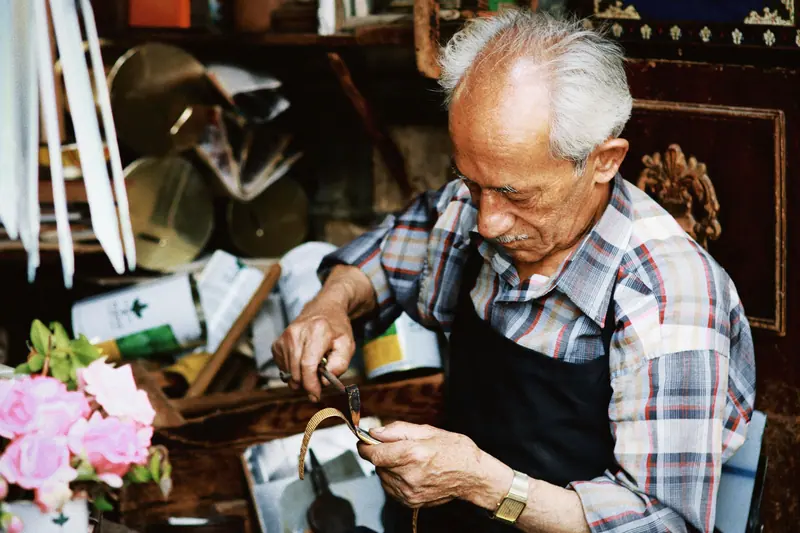 An elderly man in a workshop crafting with tools, surrounded by supplies and flowers.