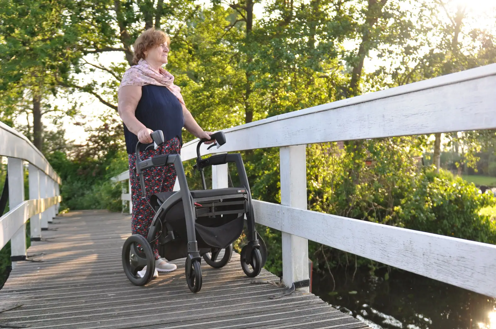 A resident with her rollator pausing on a wooden footbridge over the creek in the evening sun