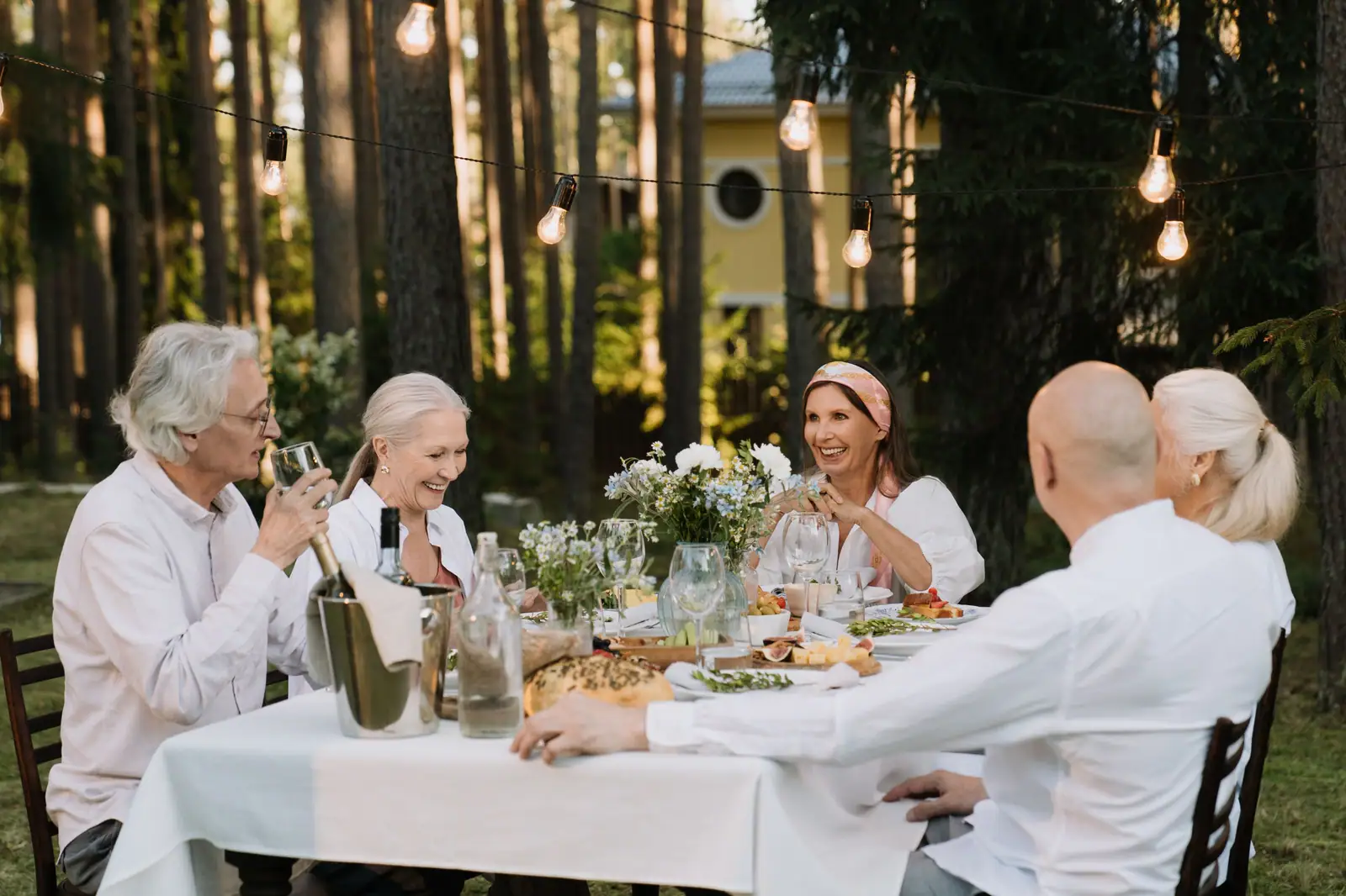 Residents laughing over supper at a long table under string lights in the pines