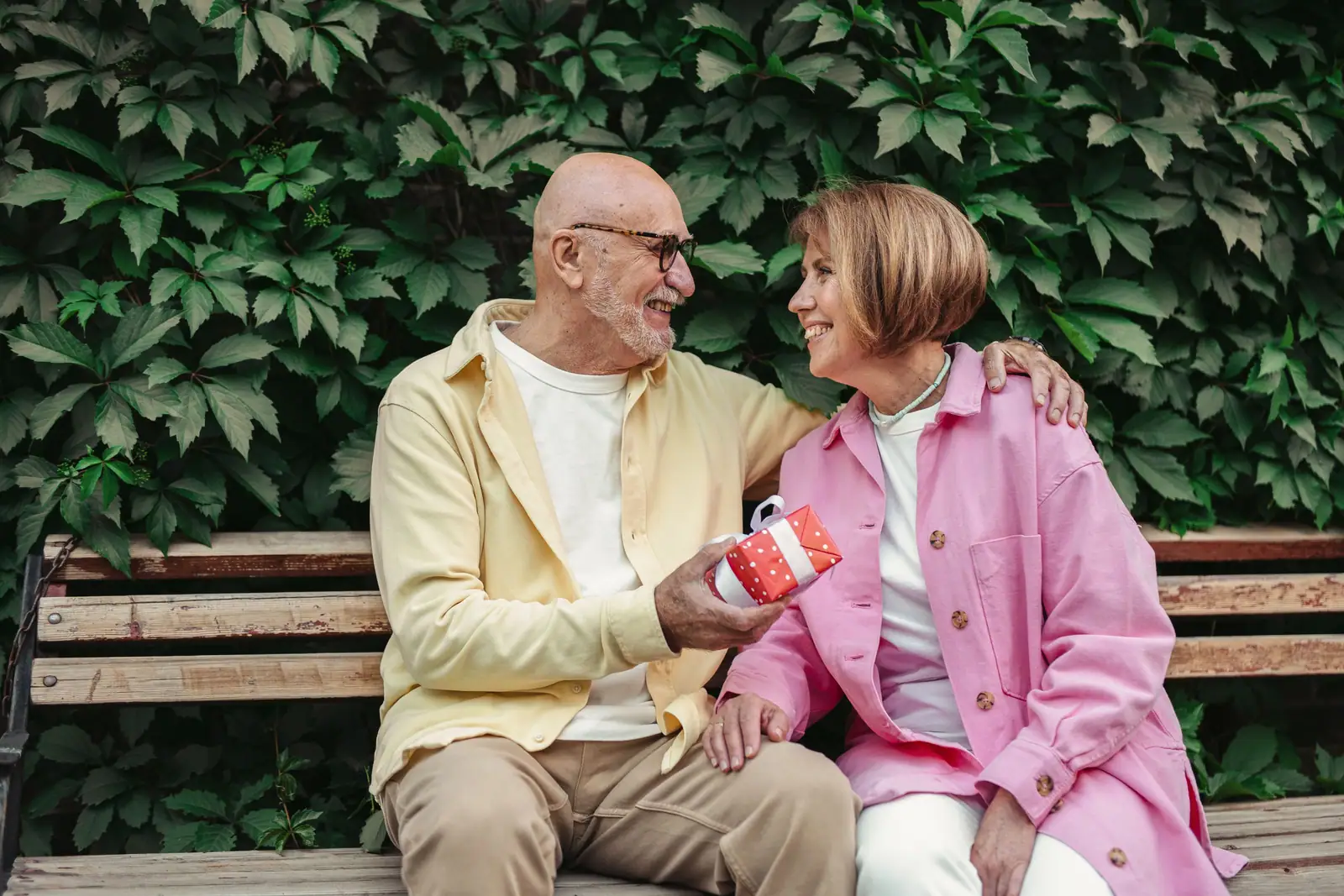 A resident in a pink jacket laughing with her husband on the garden bench, a small wrapped gift in his hand