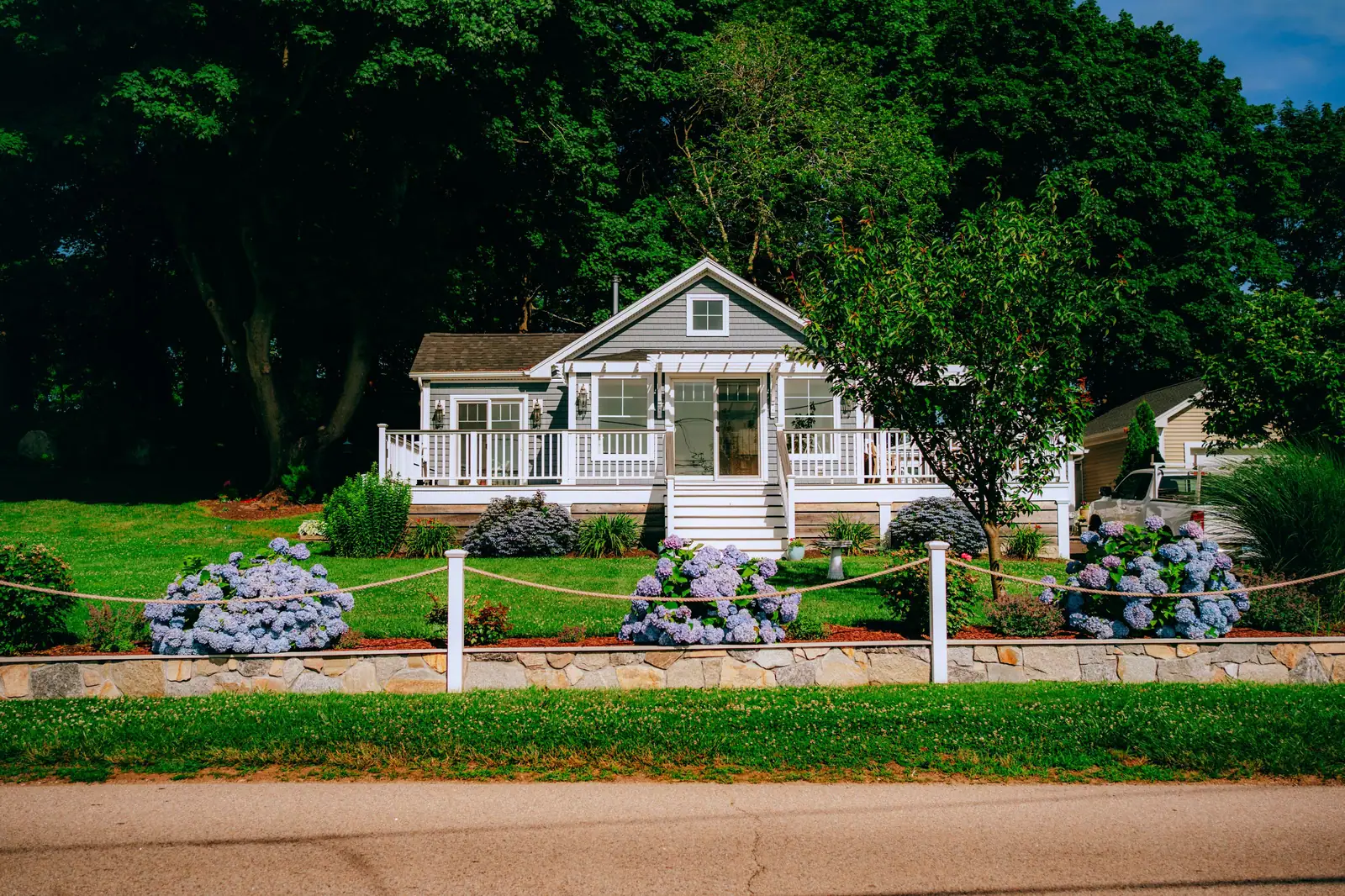 A white cottage with a wide front porch and blue hydrangeas along the walk, framed by tall oaks
