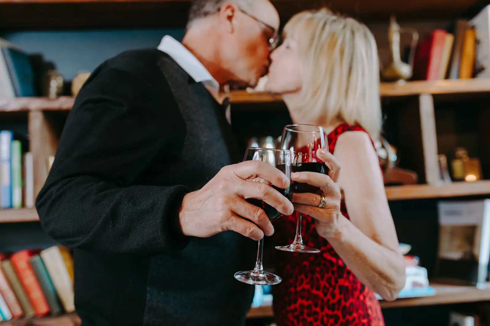 A husband and wife sharing a kiss and a glass of red wine in their apartment library