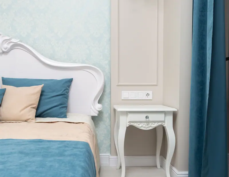 A stylish and elegant bedroom featuring a white bed, blue bedspread, and a classic nightstand.