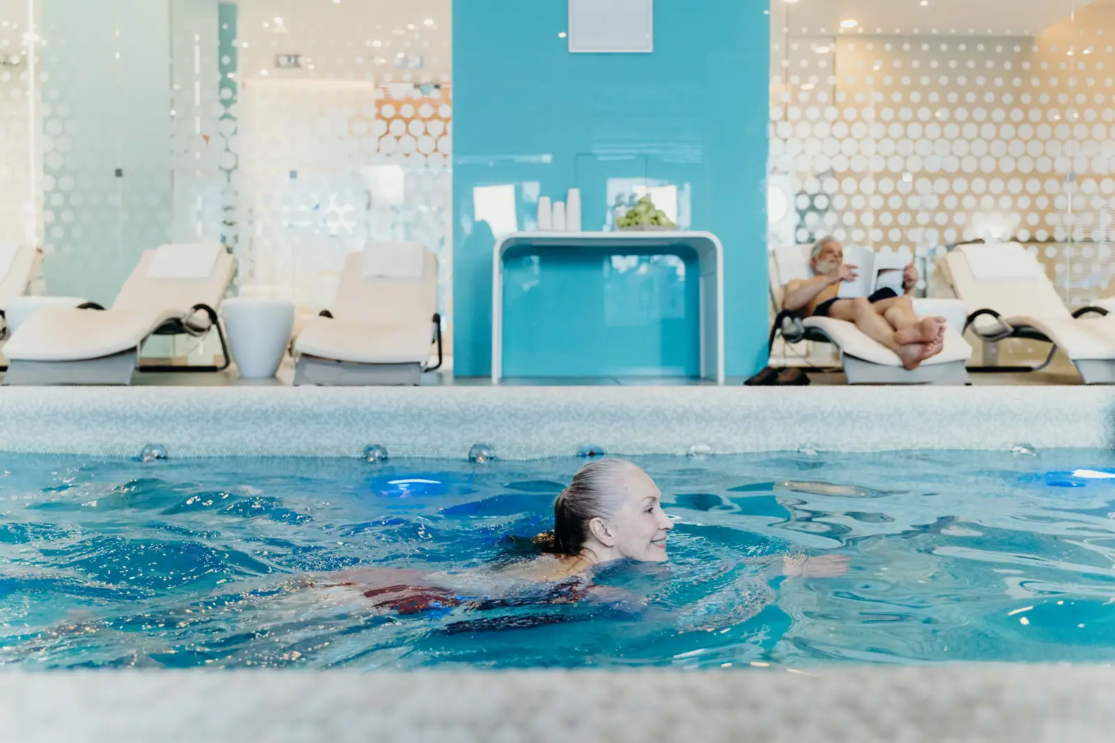 A resident swimming laps in the indoor saltwater pool while her neighbor reads on a lounge chair