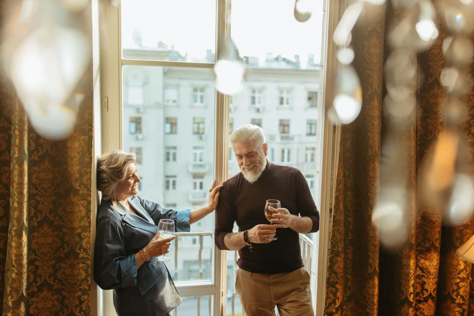 A couple sharing a glass of wine and a laugh by the tall windows of their apartment