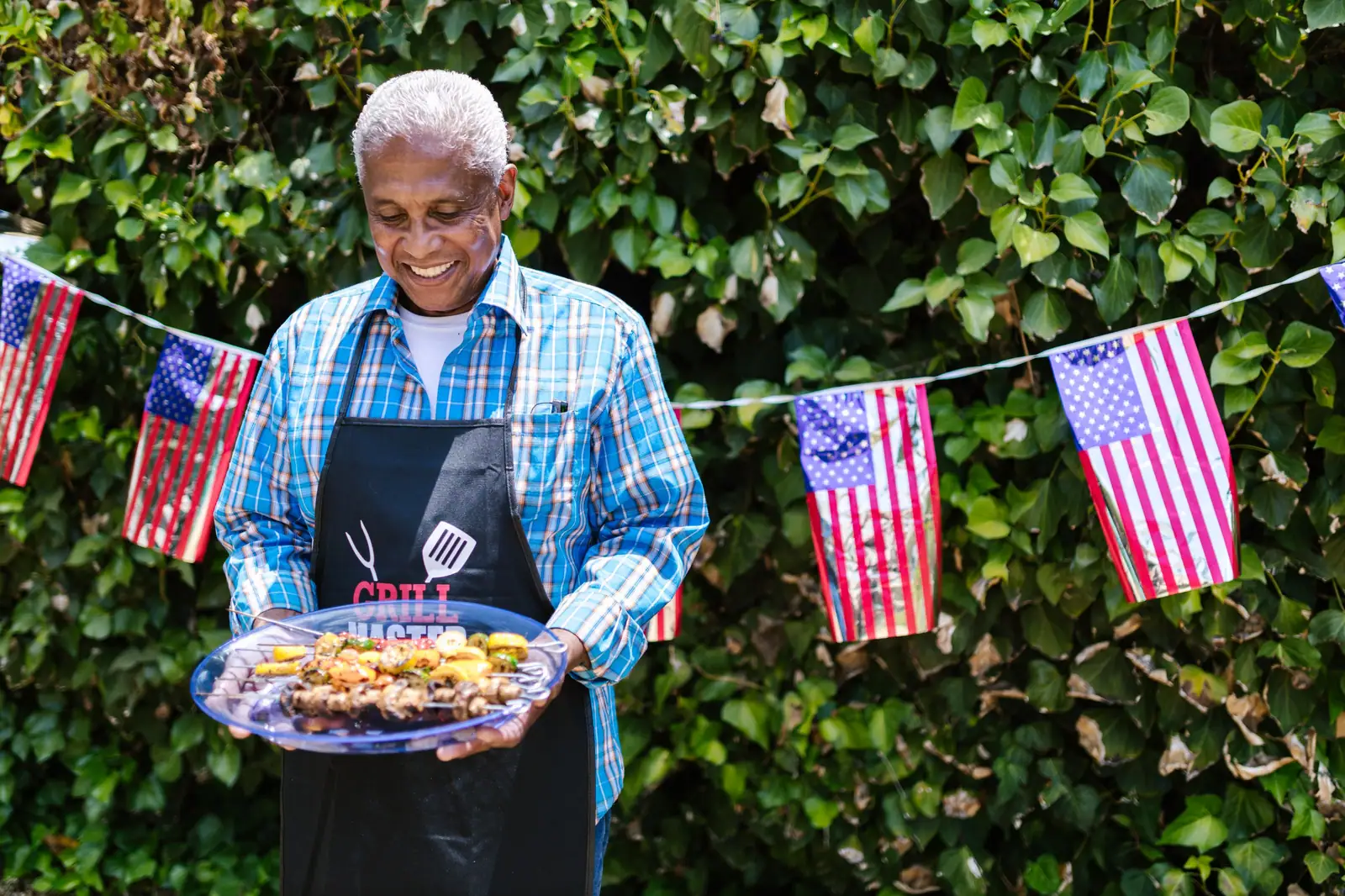 A resident in a grill-master apron proudly holding a platter of kebabs under a string of American flags