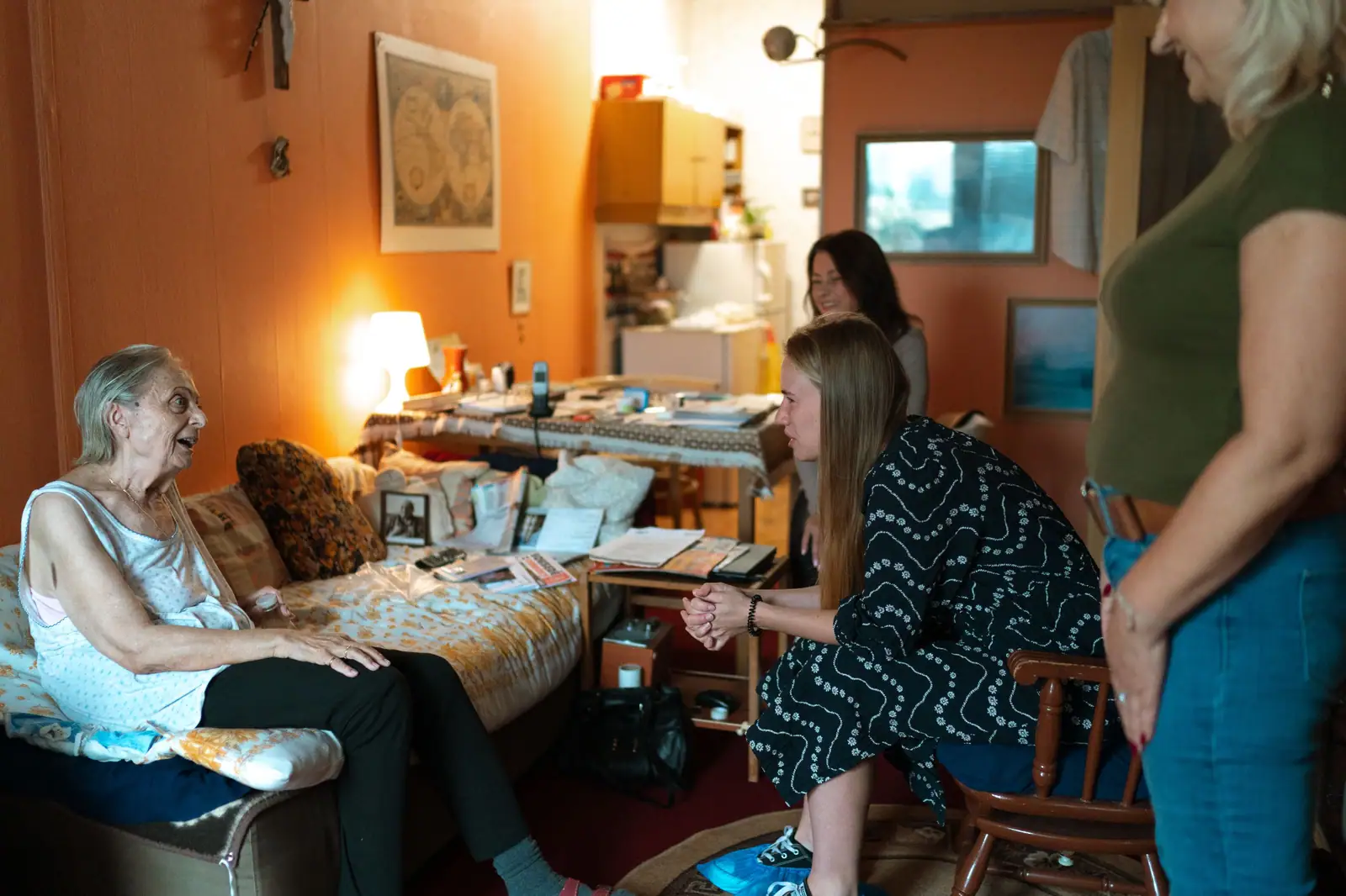 An older woman sitting in her lamp-lit living room while her daughter and a visiting nurse talk with her