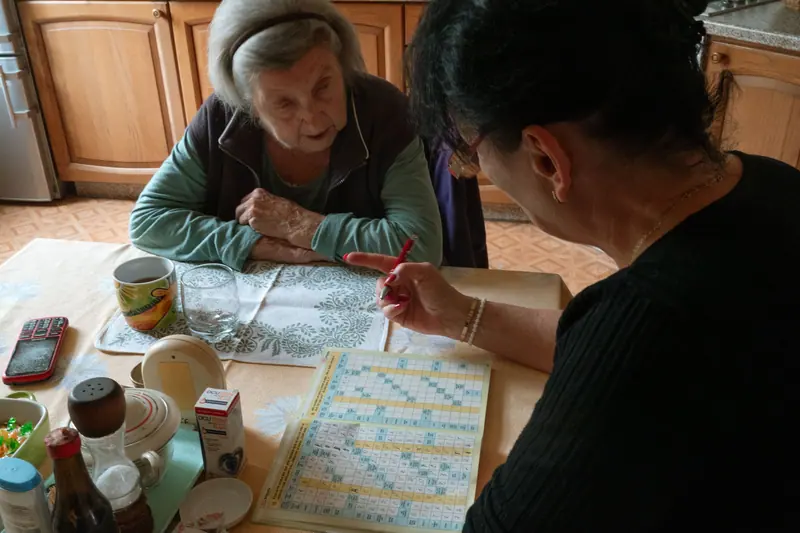 An aide and an older woman doing a crossword at the kitchen table