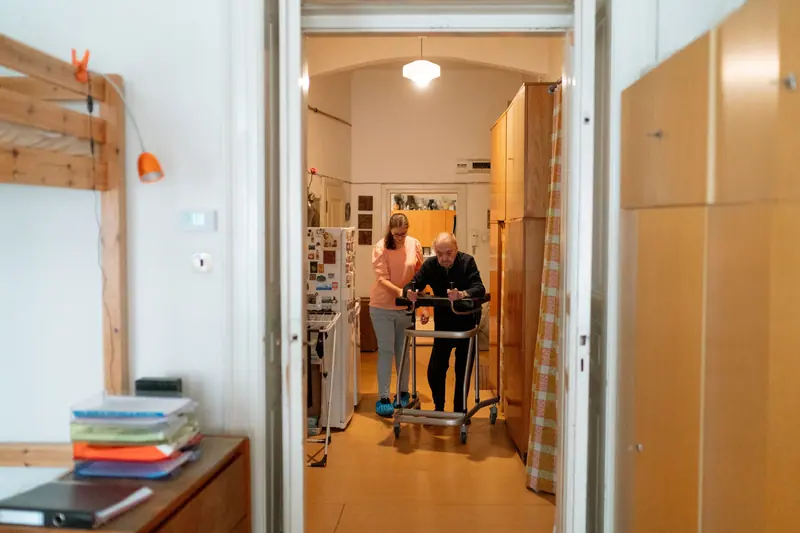 A caregiver walking beside an older man as he uses his walker down the hallway
