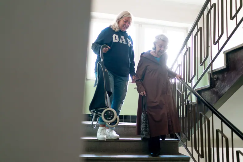 An aide walking down the stairs beside an older woman who holds the railing