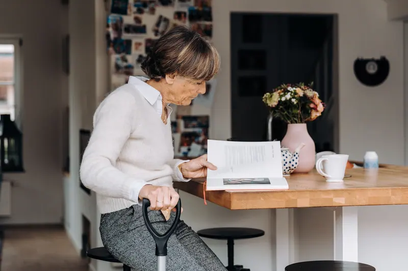 An older woman reading at her kitchen counter with her cane nearby