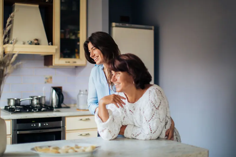 A mother and her grown daughter laughing at the kitchen counter