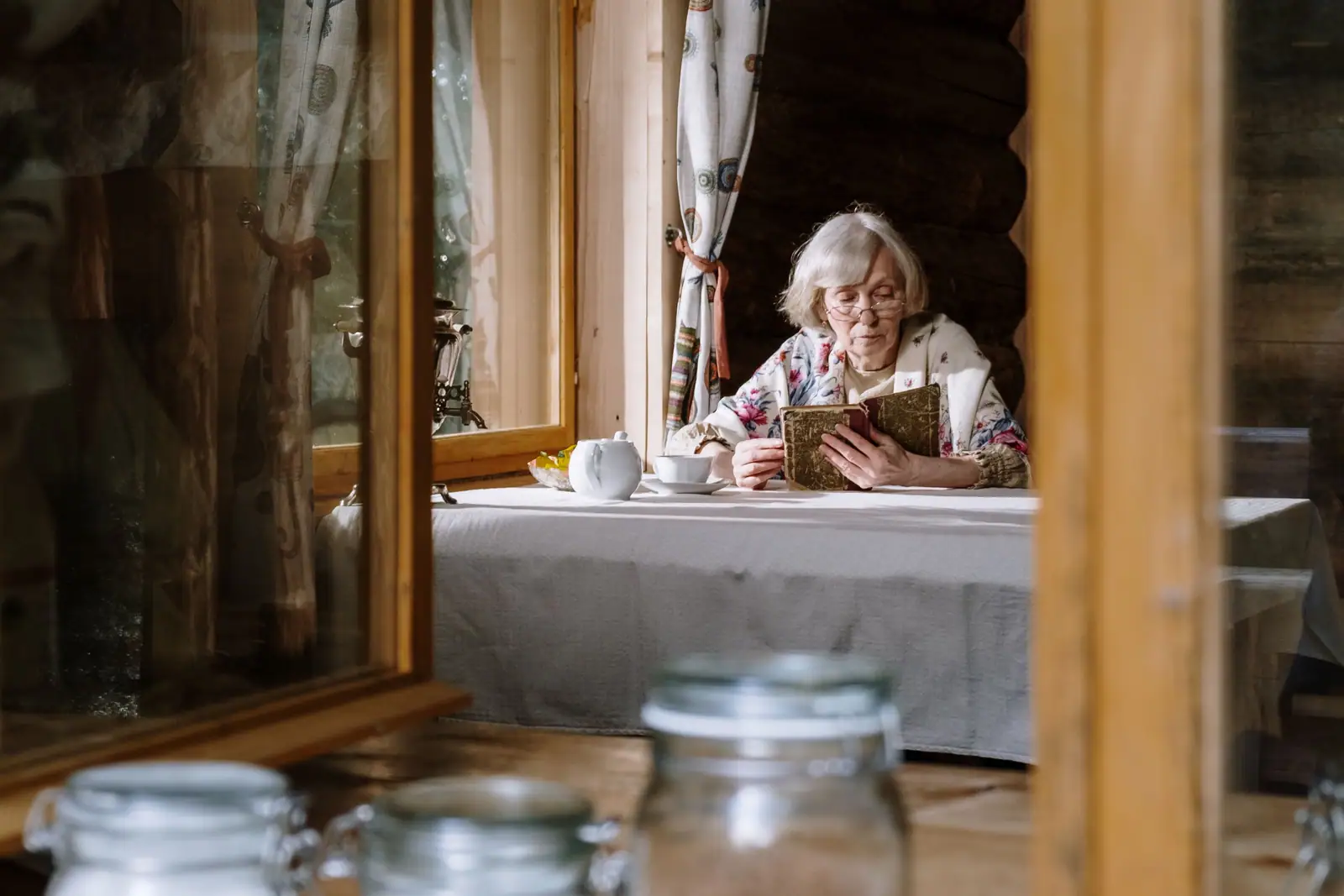 An older woman reading by a cabin window with a cup of tea, soft afternoon light on her face
