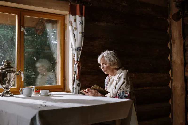 An older woman reading at a table in her cabin kitchen