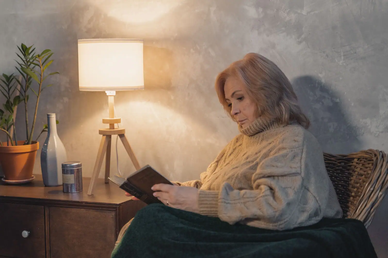 An older woman reading in a wicker armchair under a warm lamp late in the evening