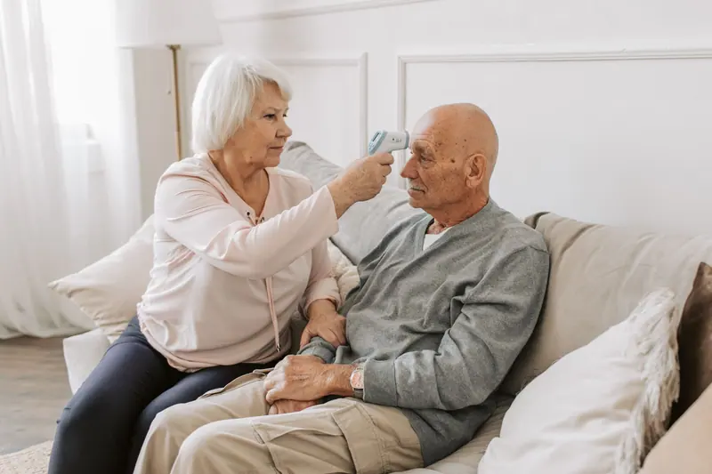 An older woman taking her husband's temperature in the living room