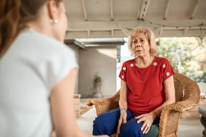 An older woman in a red blouse chatting with a visitor on her covered patio