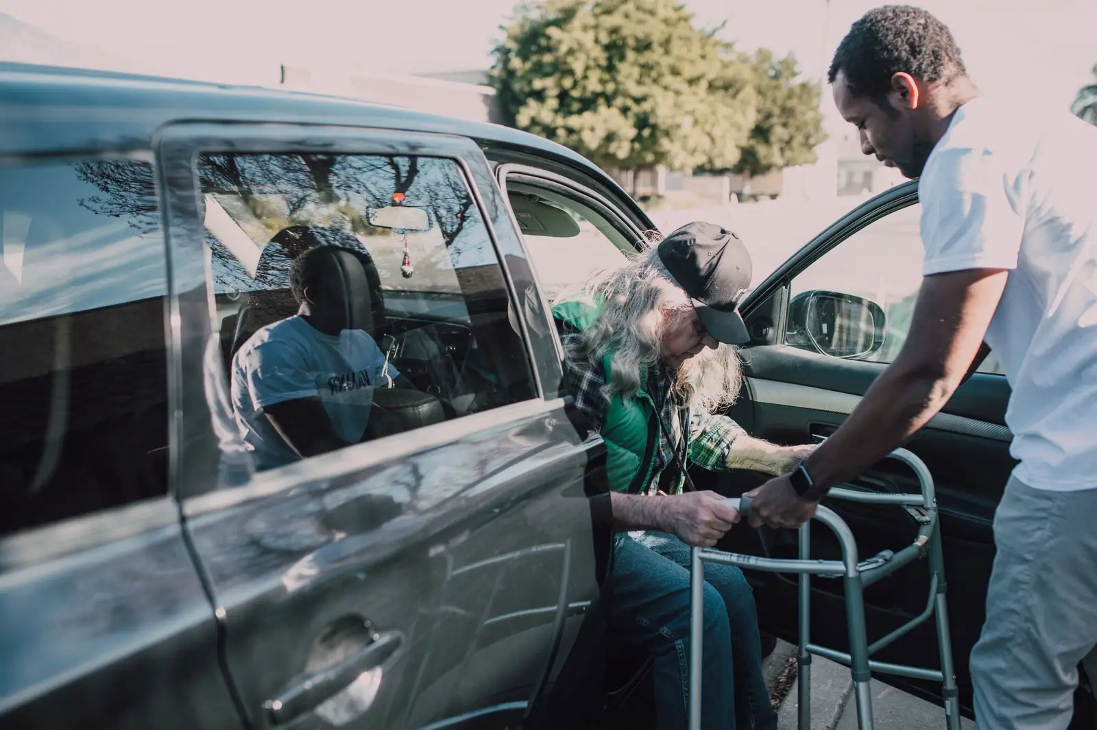 Two caregivers helping an older man with his walker as he steps out of the car at home