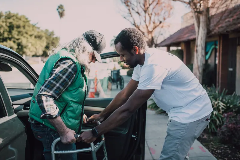 A young volunteer helping an older man with his walker by the car