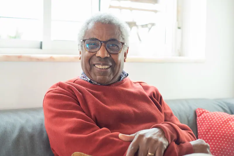 An older man in a red sweater smiling on his sofa