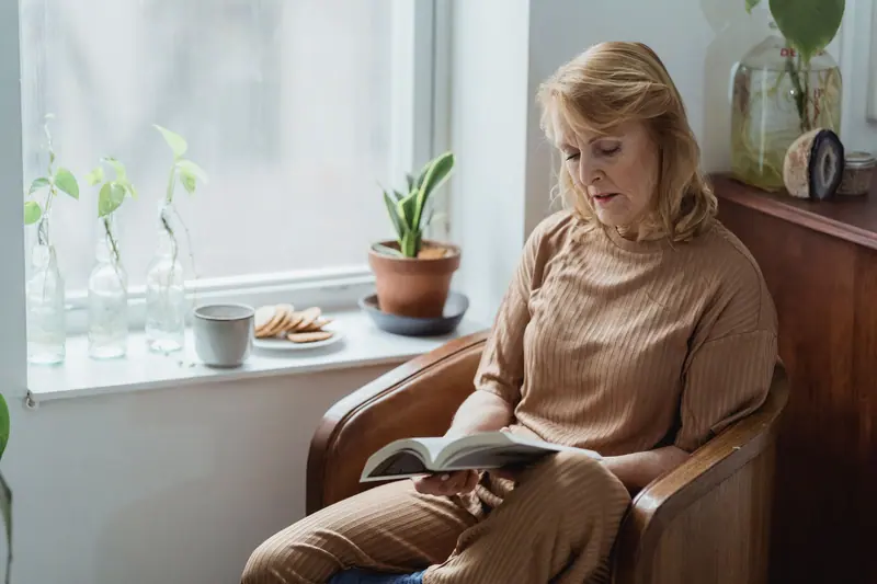 An older woman reading by the window in her armchair