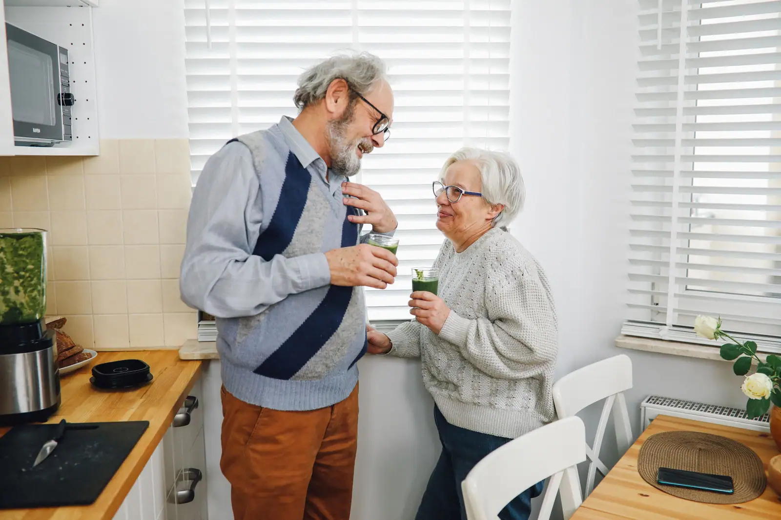 An older couple sharing a laugh over green smoothies in their sunny kitchen