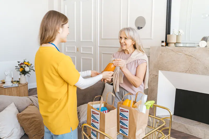An aide handing an older woman a bag of groceries in her living room