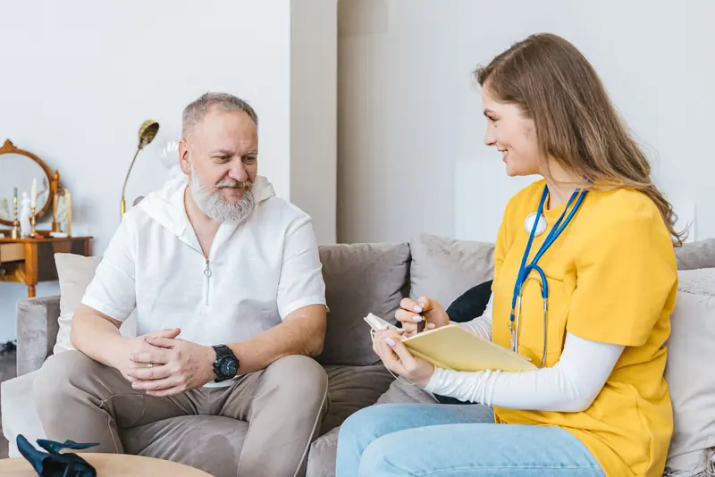 A nurse in a yellow sweater reviewing a care plan with an older man on his sofa