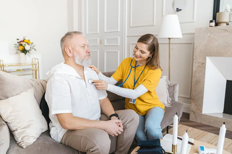 A home health nurse listening to an older man's heart on his sofa
