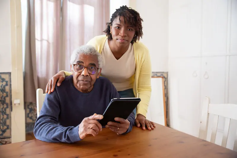 A caregiver helping an older man with his tablet at the dining table
