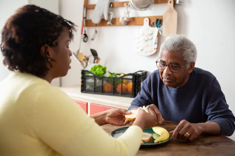 An aide sharing a plate of fruit with an older man in his kitchen