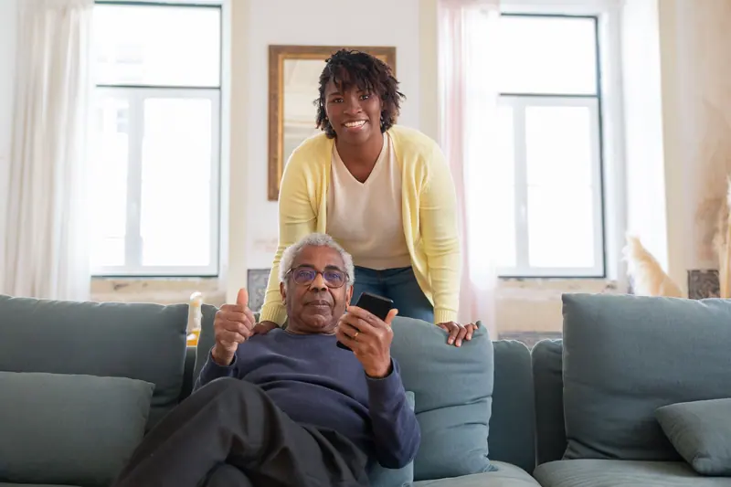 An older man laughing on his sofa as his nurse leans in