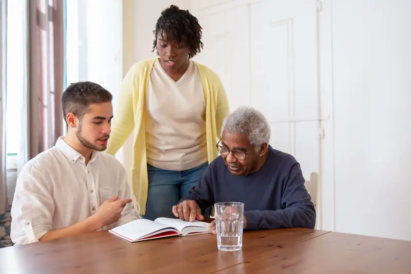 Two caregivers helping an older man with his notebook at the table