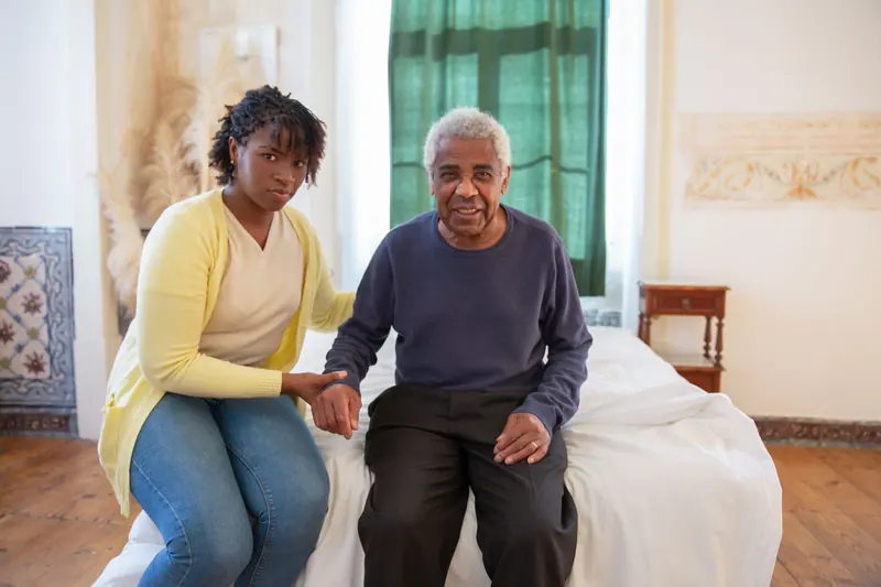 A hospice aide sitting beside a patient on the edge of his bed