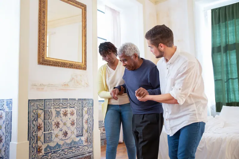 Two caregivers steadying an older man as he walks across his bedroom