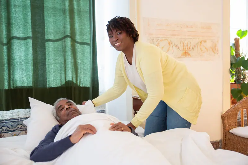 A nurse tucking a blanket around an older man resting in his bedroom