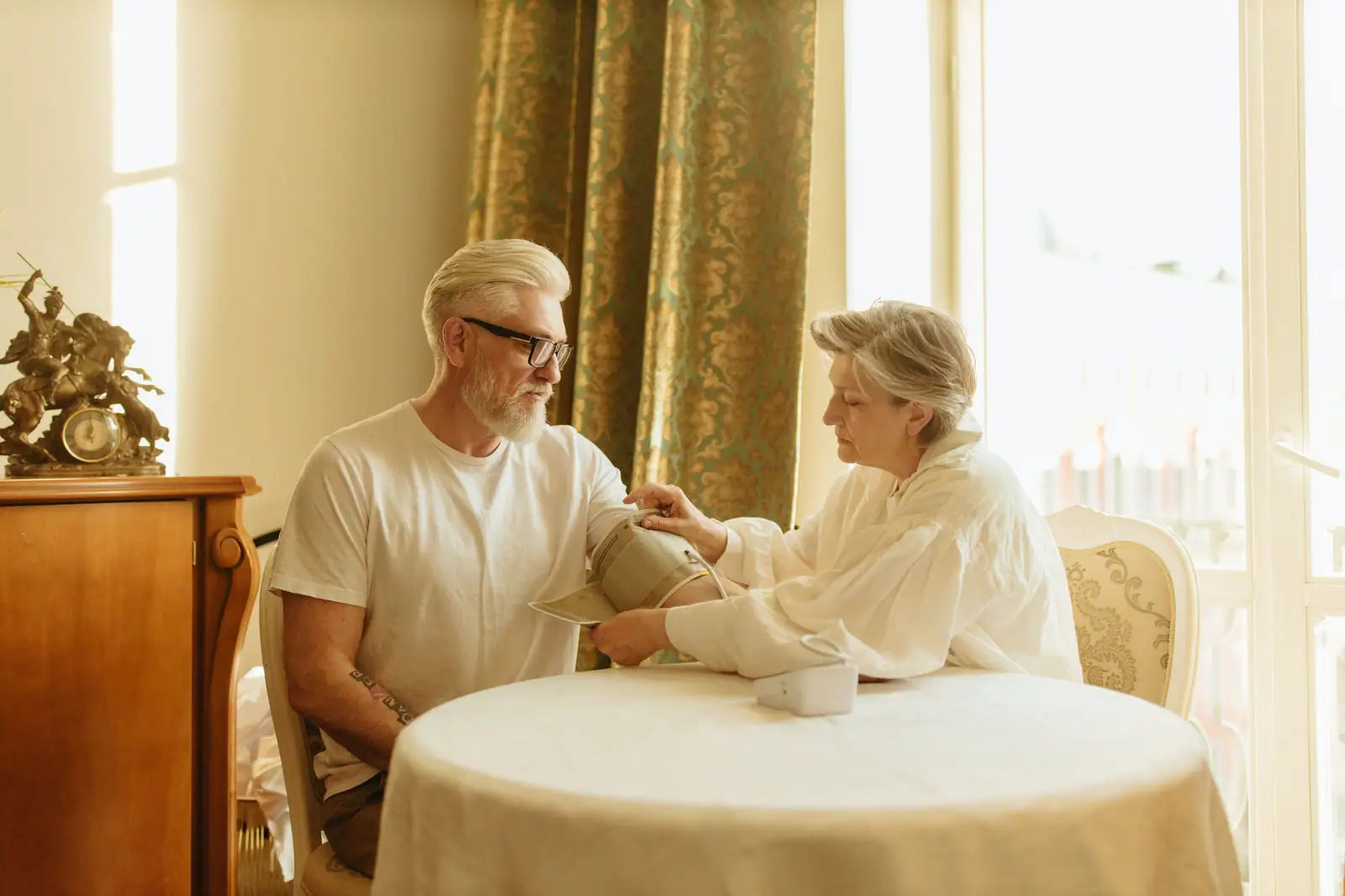 An older couple checking blood pressure together at a round table by sunlit gold curtains