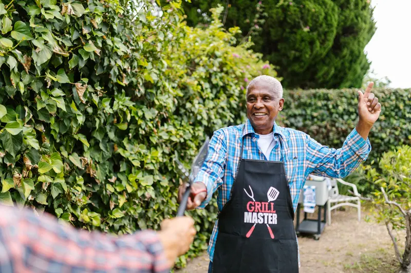 An older man in a grilling apron waving a spatula in his backyard