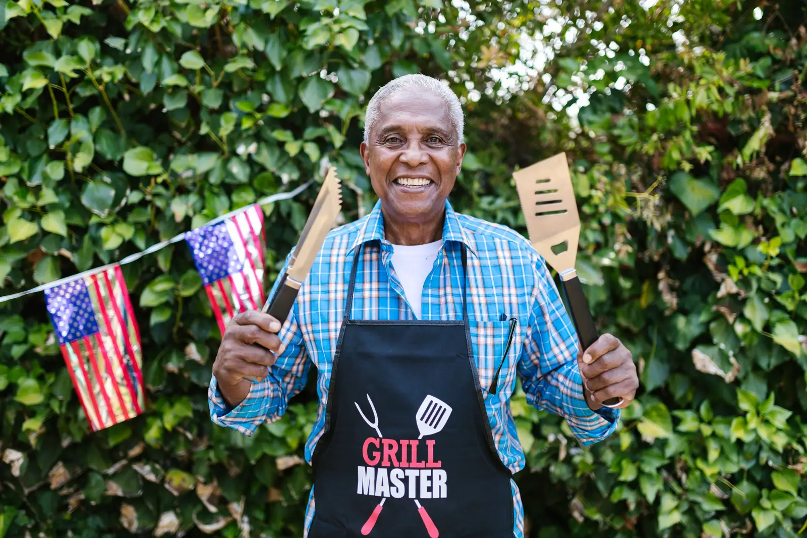 A smiling resident in a Grill Master apron holding a spatula and a little flag in front of the patio hedge