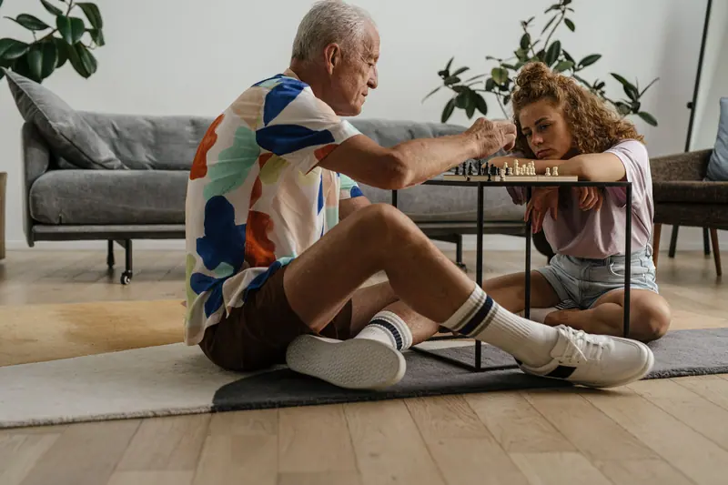 An older man and his granddaughter playing chess on the living-room floor
