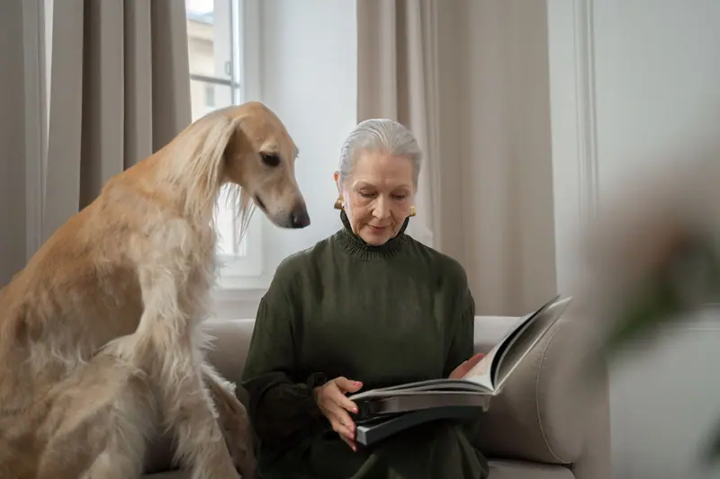 A woman reading on the couch as her dog leans in