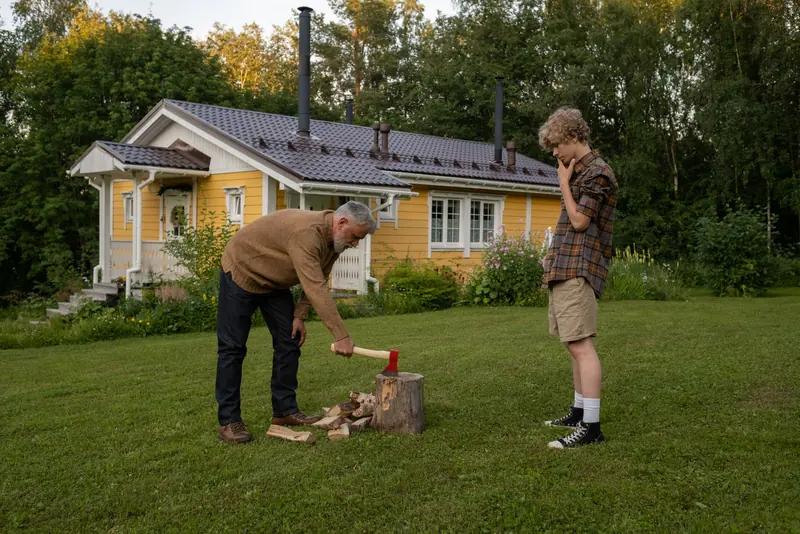 A grandfather splitting firewood in the yard while his grandson watches
