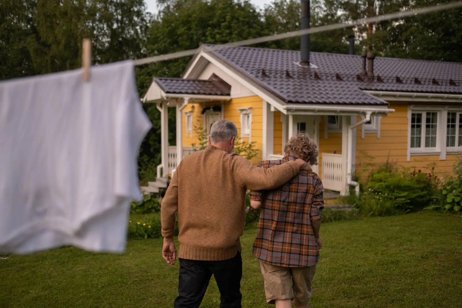 An older couple walking arm in arm across the grass toward their yellow farmhouse, laundry on the line