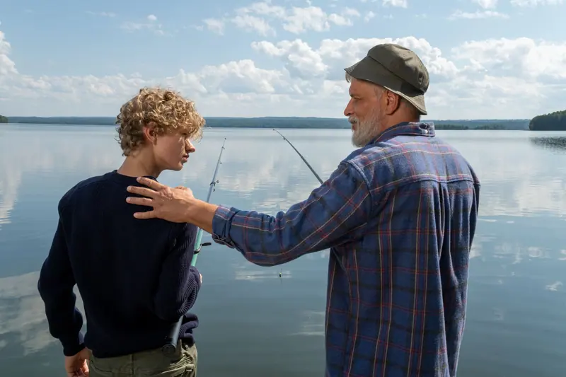 A grandfather resting his hand on his grandson's shoulder at the water's edge