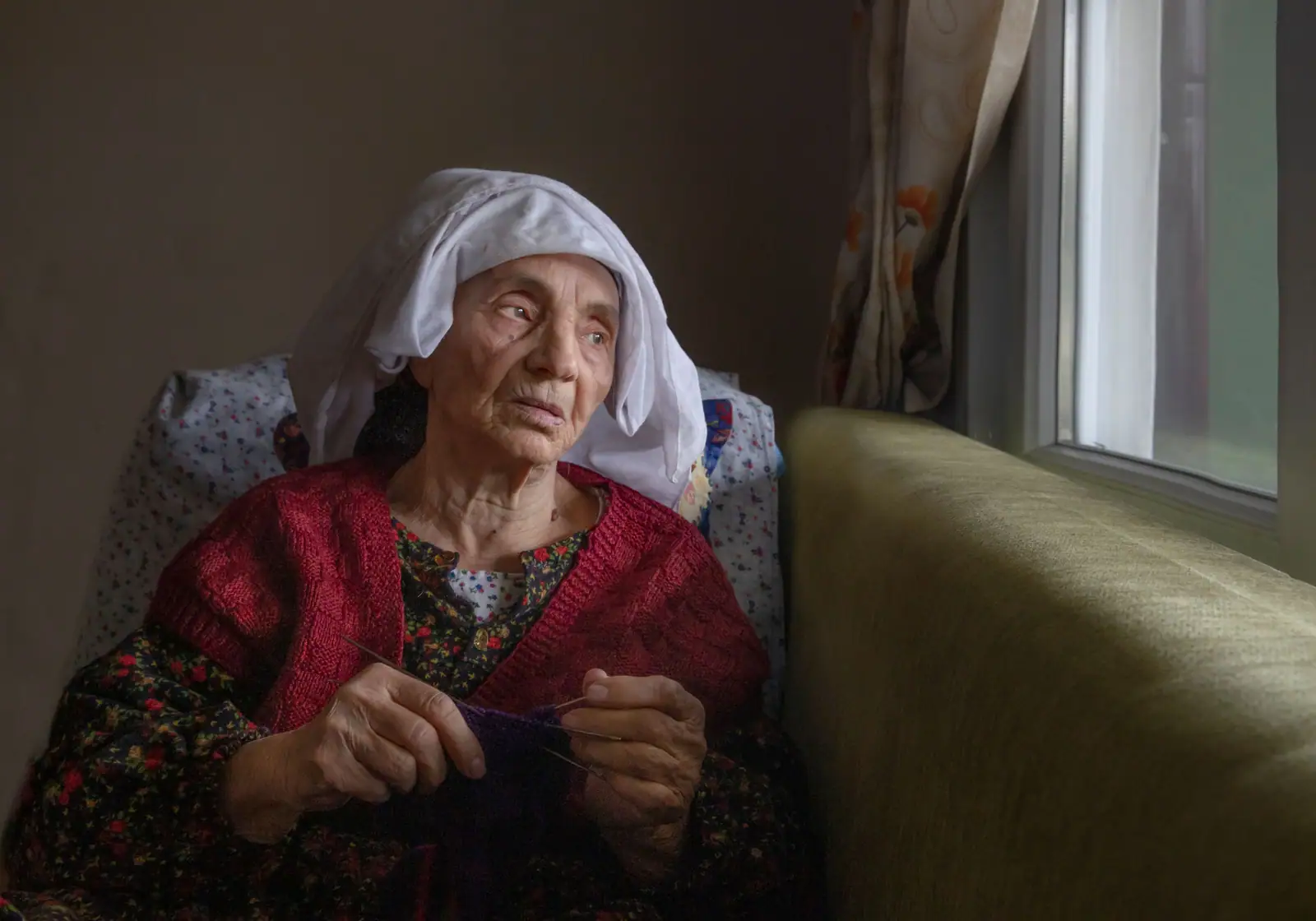 A resident in a white headscarf and red embroidered vest sitting in the lamplight of her room, gazing toward the window