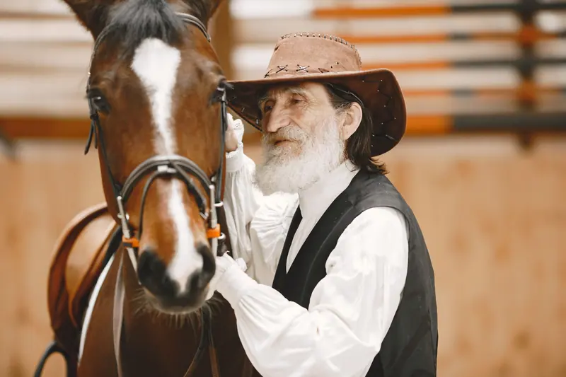 An old horseman in a felt hat with his bay horse in the barn