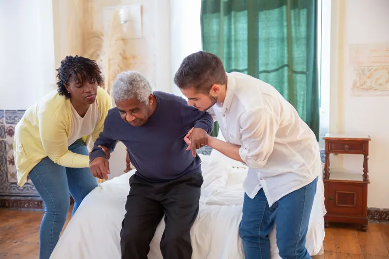 Two caregivers helping a patient stand from the edge of his bed