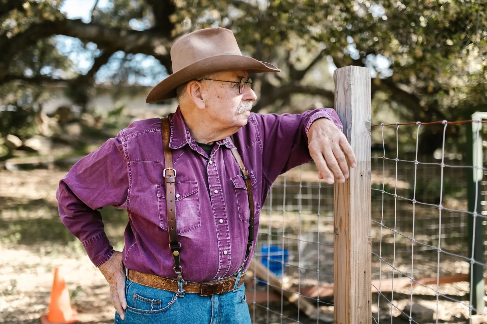 A rancher in a felt hat and purple work shirt leaning on his fence post under the oaks