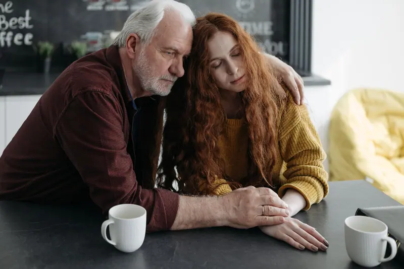 A daughter resting her head on her father's shoulder at the kitchen table